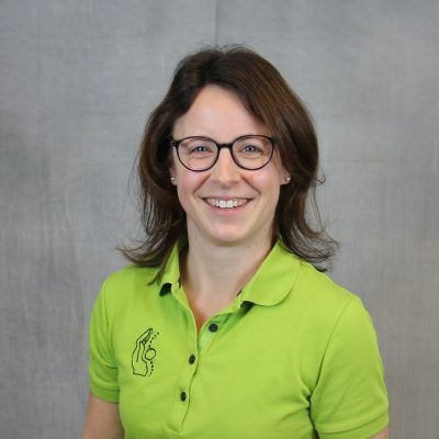 Porträt einer lächelnden Mitarbeiterin des Osteopathiezentrums in grünem Poloshirt vor hellem Hintergrund.