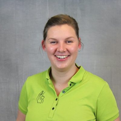 Porträt einer lächelnden Mitarbeiterin des Osteopathiezentrums in grünem Poloshirt vor hellem Hintergrund.