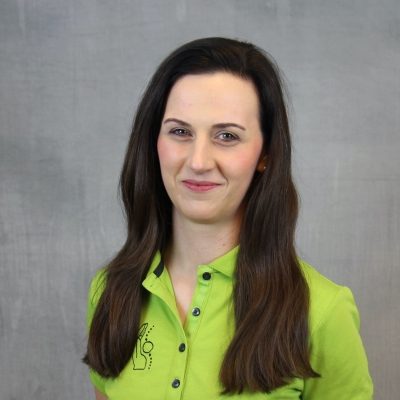Porträt einer lächelnden Mitarbeiterin des Osteopathiezentrums in grünem Poloshirt vor hellem Hintergrund.