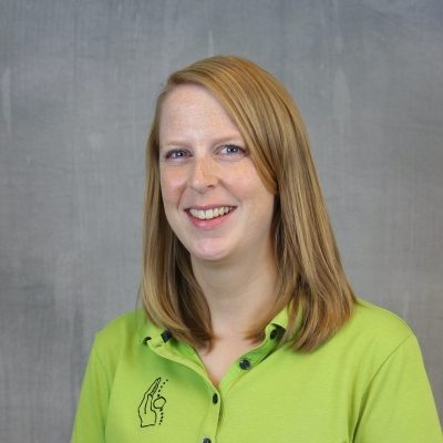 Porträt einer lächelnden Mitarbeiterin des Osteopathiezentrums in grünem Poloshirt vor hellem Hintergrund.