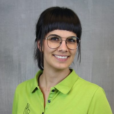 Porträt einer lächelnden Mitarbeiterin des Osteopathiezentrums in grünem Poloshirt vor hellem Hintergrund.