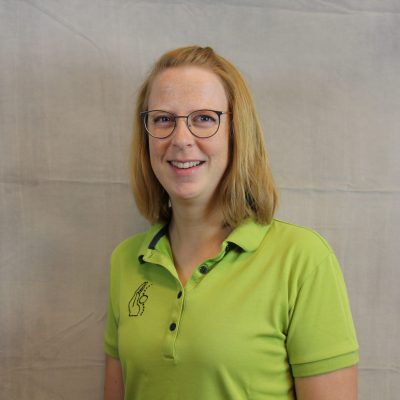 Porträt einer lächelnden Mitarbeiterin des Osteopathiezentrums in grünem Poloshirt vor hellem Hintergrund.