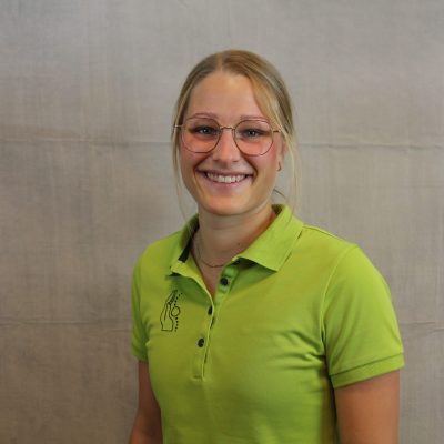 Porträt einer lächelnden Mitarbeiterin des Osteopathiezentrums in grünem Poloshirt vor hellem Hintergrund.