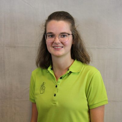 Porträt einer lächelnden Mitarbeiterin des Osteopathiezentrums in grünem Poloshirt vor hellem Hintergrund.
