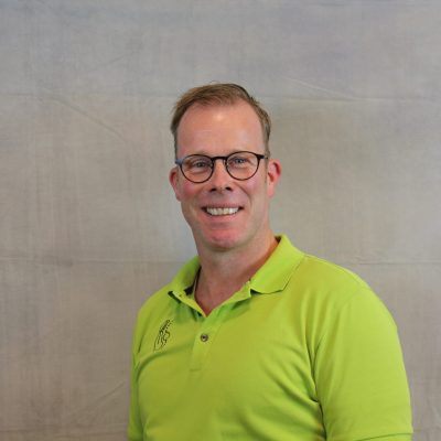 Porträt eines lächelnden Mitarbeiters mit Brille und Bart in grünem Poloshirt vor hellem Hintergrund.