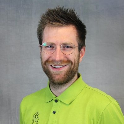 Porträt eines lächelnden Mitarbeiters mit Brille und Bart in grünem Poloshirt vor hellem Hintergrund.