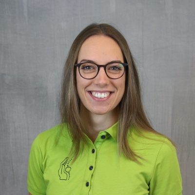 Porträt einer lächelnden Mitarbeiterin des Osteopathiezentrums in grünem Poloshirt vor hellem Hintergrund.