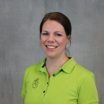 Porträt einer lächelnden Mitarbeiterin des Osteopathiezentrums in grünem Poloshirt vor hellem Hintergrund.