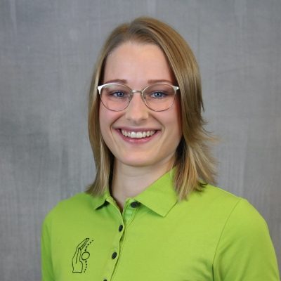 Porträt einer lächelnden Mitarbeiterin des Osteopathiezentrums in grünem Poloshirt vor hellem Hintergrund.