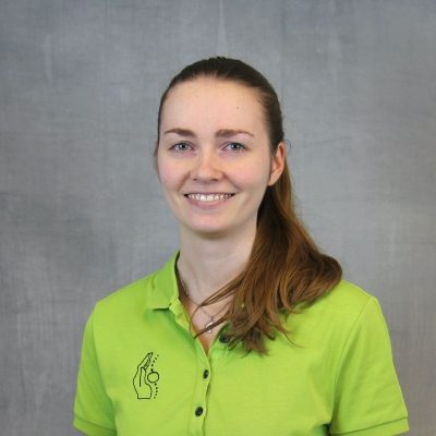 Porträt einer lächelnden Mitarbeiterin des Osteopathiezentrums in grünem Poloshirt vor hellem Hintergrund.