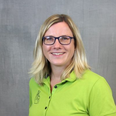 Porträt einer lächelnden Mitarbeiterin des Osteopathiezentrums in grünem Poloshirt vor hellem Hintergrund.